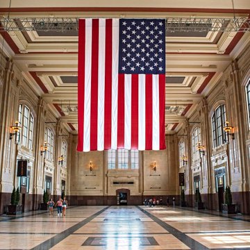 A massive American flag hangs prominently from the ceiling of an ornate government-style hall with marble floors and arched windows. The image's setting in what appears to be a U.S. government building visually reinforces the post's news about US government-linked wallets moving approximately $1 billion in Bitcoin from a Bitfinex hack seizure wallet.