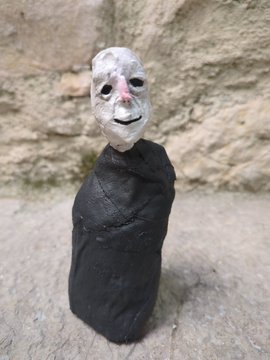 A small handcrafted figurine with a white clay smiling face draped in dark fabric stands against a rough stone background.