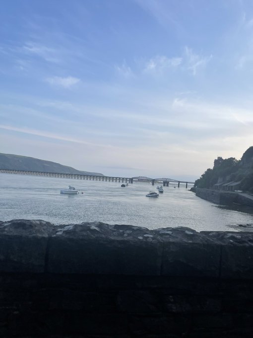 A calm morning view of a quiet bay with small boats anchored beneath a distant bridge, framed by a stone wall in the foreground, reflecting the simple, peaceful beauty the message celebrates.