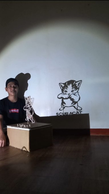 A young man sits beside a cardboard box with "SCUB-A" written on it, featuring a delicate cutout shadow art sculpture of a kitten, echoing the "SCUBACAT" logo projected on the wall behind him—a six-hour handmade tribute to the $SCUBACAT community.