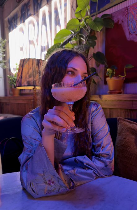 A portrait of a young woman with long dark hair holding and sipping a pink cocktail from a coupe glass through a black straw, set in a dimly lit venue with neon lights and plants in the background.