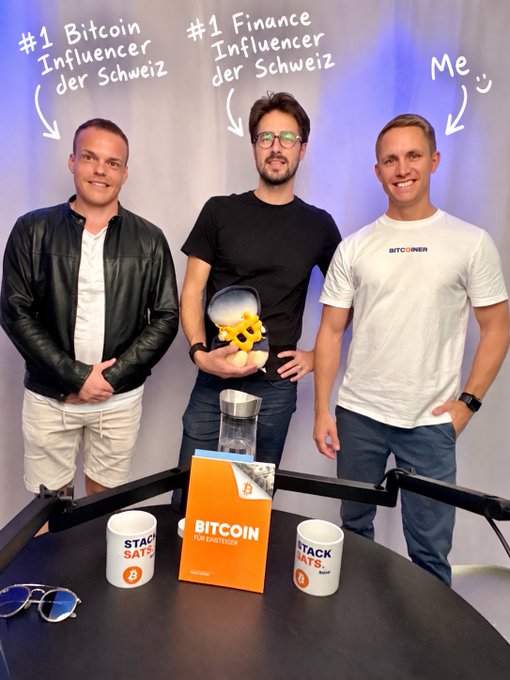Three men standing together in a studio setting, labeled with text as '#1 Bitcoin Influencer der Schweiz,' '#1 Finance Influencer der Schweiz,' and 'Me,' with Bitcoin-themed merchandise on a table in front of them.