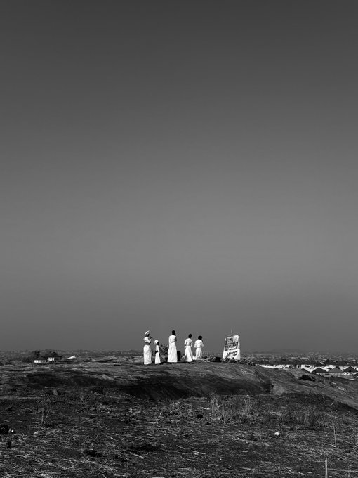 A black-and-white photograph captures a group of people in traditional attire standing on a rocky hilltop under a vast, dark sky.