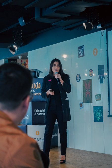 The image shows a woman standing at a podium, speaking into a microphone while wearing a black jacket. Behind her, there are several posters displayed on the wall, including one that reads "Zcon Vozes 2". The background of the image is blurred, but it appears to be an indoor setting with a blue ceiling and white walls.