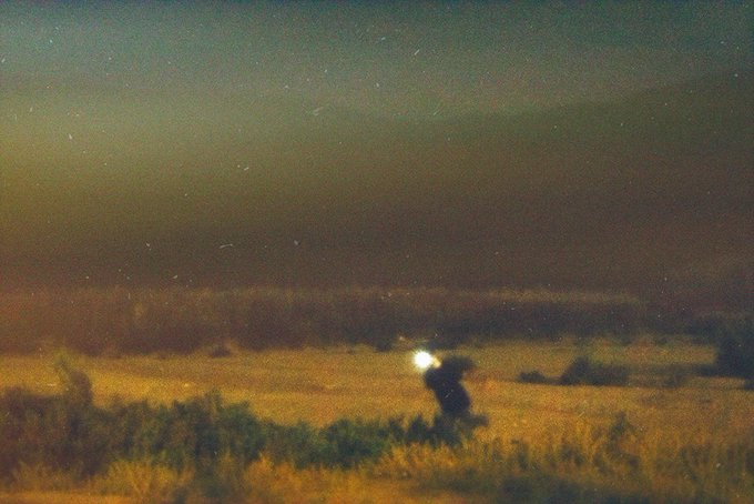 A blurry night photograph shows a person standing in a field with a light source, set against a dark sky with visible stars and a tree line on the horizon.