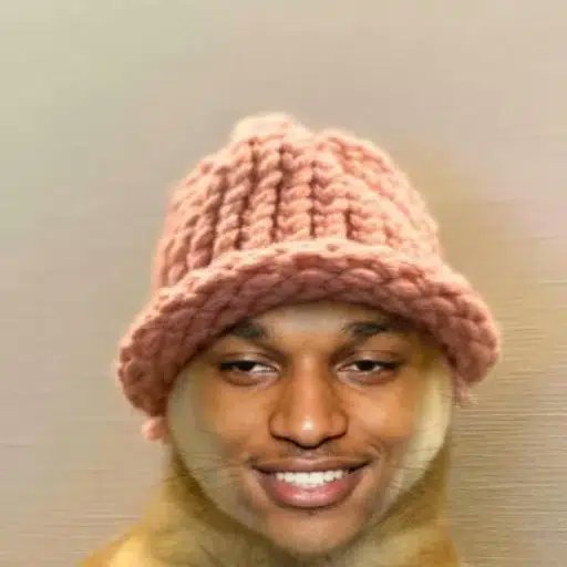A smiling young man wearing a pink knitted bucket hat, photographed against a plain gray background.