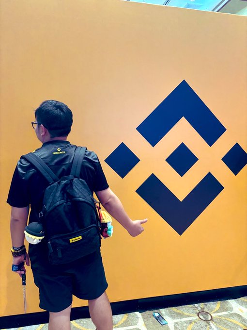 A man wearing glasses, black shorts, and a backpack with a 'BINANCE' logo points at a large blue Binance logo on a yellow wall.