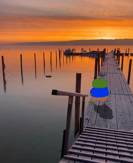 A scenic sunset over a calm lake featuring a wooden dock with a cartoon frog character (resembling Pepe) in a blue shirt standing on the dock, set against an orange sky.