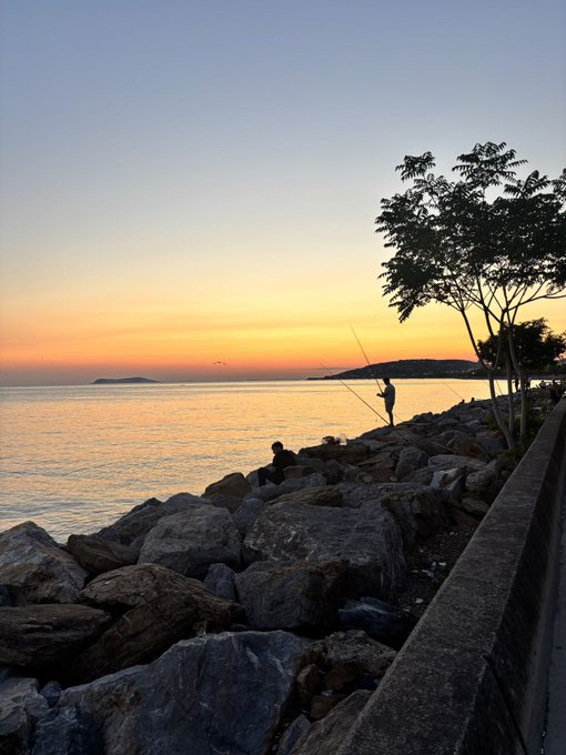 A serene sunset scene over a calm body of water, with silhouettes of people fishing on rocky shoreline and a tree visible on the right.