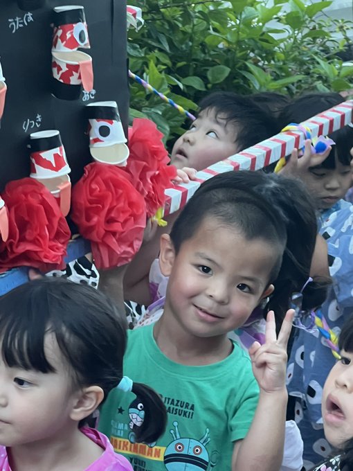 A group of young Japanese children pose together, with one boy in a green Ultraman/Kitaro shirt making a peace sign, while others hold craft decorations resembling sushi rolls and red paper flowers.