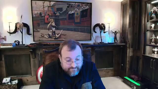 The image shows a man sitting at a desk with a computer monitor in front of him. He has glasses on his face and appears to be focused on something on the screen. The background features a painting hanging on the wall, adding an artistic touch to the room. There are also two lamps present in the scene, one located near the center of the image and another towards the right side.