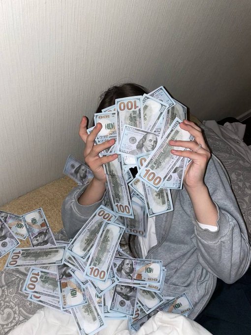 A person holds fanned-out stacks of $100 bills in front of their face while sitting on a bed scattered with more $100 bills, visually representing the promise of crypto wealth and millionaire success. The image's display of large amounts of cash aligns with the post's bold claim about becoming a millionaire through cryptocurrency.
