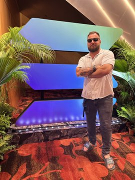 A bearded man in a white shirt and sunglasses stands confidently with arms crossed in front of a glowing blue digital display framed by tropical plants, at what appears to be a Solana-sponsored event. The image complements the post by visually representing the speaker's dedicated involvement with the Solana ecosystem, reinforcing the message of long-term commitment and belief in the blockchain's builder community.
