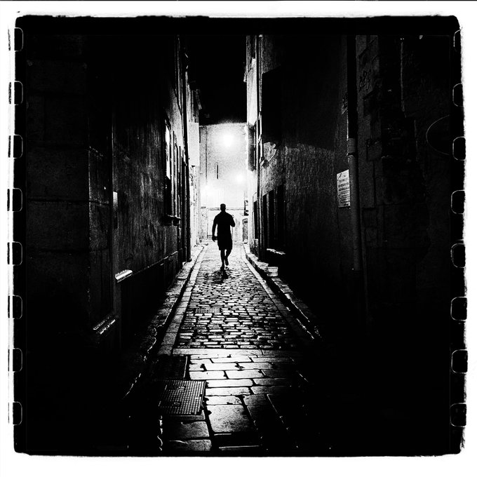 A black and white photograph of a person walking alone down a narrow, cobblestone alleyway at night, lit by a distant light source, with a film border frame.