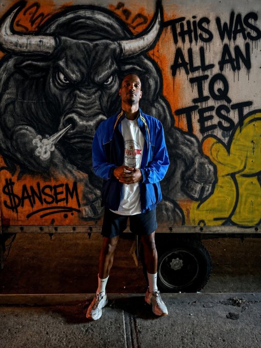 A man in a blue jacket and white t-shirt stands in front of a graffiti mural depicting a bull with the text 'THIS WAS ALL AN IQ TEST' and '$ANSEM' tagged on it.