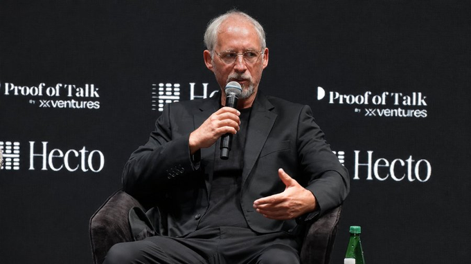 The image features a man sitting in a chair with his hands outstretched as if he's giving a speech or presentation. The background of the image shows a black wall with white text, which includes the words "proofoftalk", "x ventures", and "bullish". The man is wearing a suit and tie, suggesting that this event might be related to a formal gathering or conference.