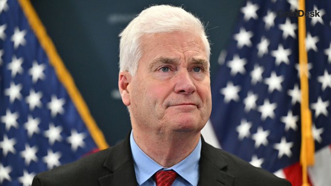 An older man with white hair in a dark suit and red tie stands before American flags, with a 'News Desk' watermark in the corner.