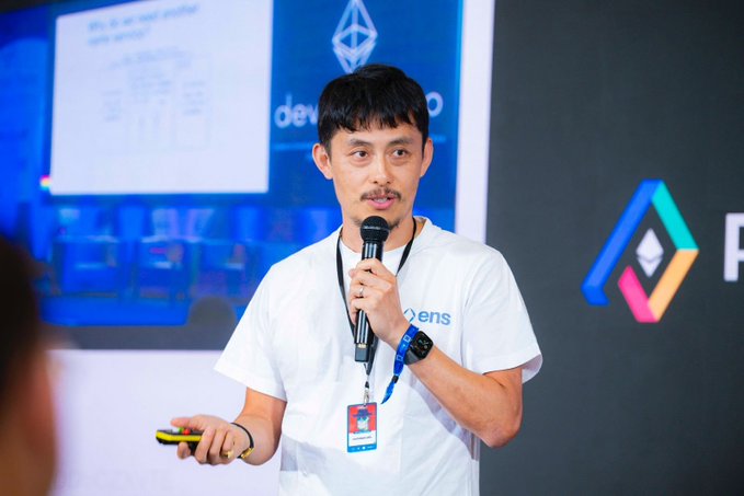 Makoto Inoue, wearing a white ENS t-shirt and lanyard, speaks into a microphone while presenting on stage at Pragma Lisbon, with an "ens" logo and presentation slides visible in the background.