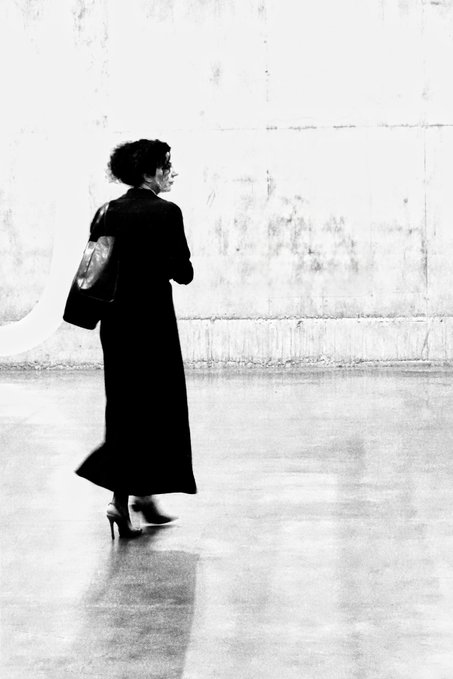 A black and white photograph of a woman in a long dark coat walking with a handbag, set against a minimalist concrete background.