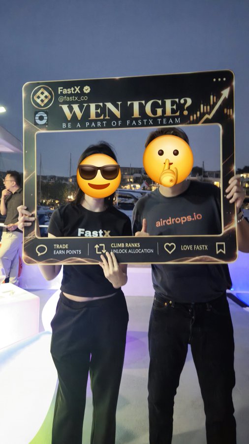 Two people stand outdoors at dusk holding a large photo frame that reads 'FastX @fastx_co WEN TGE? BE A PART OF FASTX TEAM'. The individuals' faces are obscured by emoji stickers, and one wears a shirt with 'airdrops.io'.