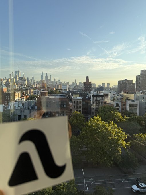 A view through a window showing a city skyline at dusk, with a paper featuring a stylized 'a' logo taped to the glass in the foreground.