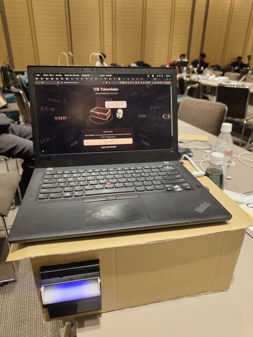 A black ThinkPad laptop on top of a cardboard box, displaying a login screen for "SE Takarazuka" with a card reader device positioned underneath the box.