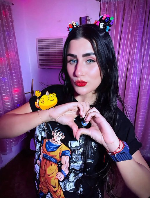 A woman wearing a Dragon Ball Z Goku t-shirt and pixel-art character hair clips, forming a heart shape with her hands, in a room with purple lighting and curtains.