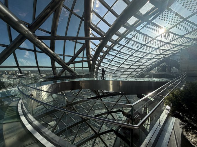 An interior view of a modern glass and steel architectural structure with a spiral walkway, featuring a person standing by the railing overlooking the lower levels.