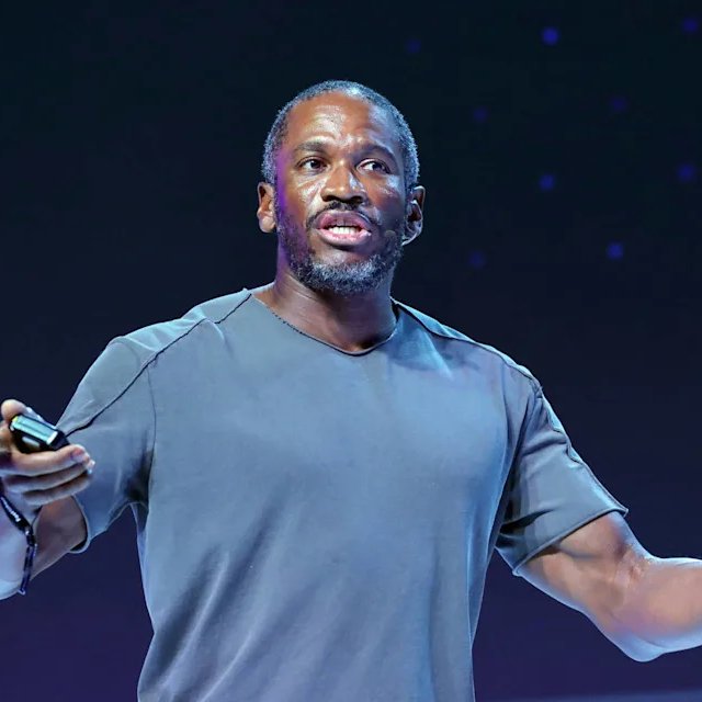 A Black man in a gray t-shirt holds a small device while speaking on a dark stage with starry lights behind him.