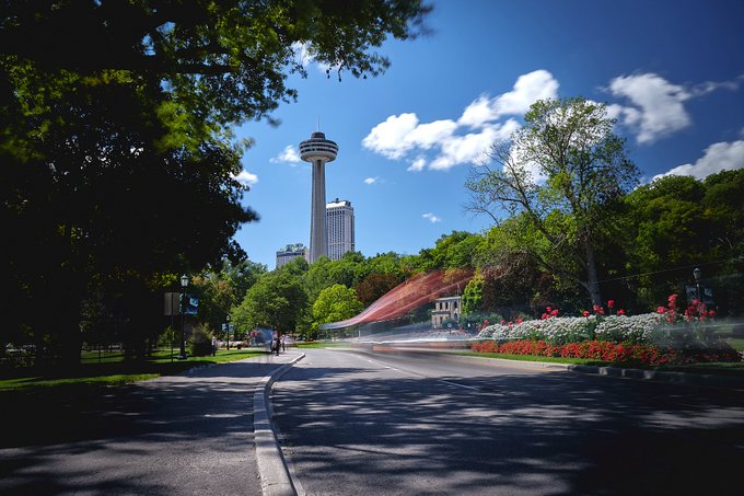 A scenic outdoor photograph showing a curving paved road lined with lush green trees and flowers, with the Skylon Tower visible in the distance against a blue sky with scattered clouds.