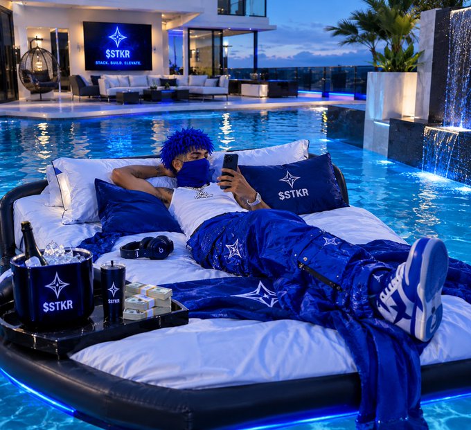 A person with blue hair lies on a pool float branded with the '$STKR STACK. BUILD. ELEVATE.' logo, holding a phone, surrounded by matching branded accessories and stacks of cash.