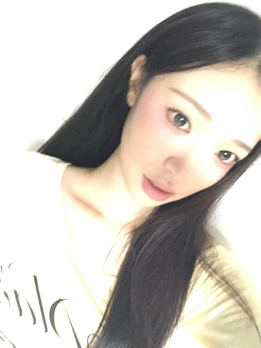 A close-up portrait of a woman with long black hair wearing a yellow top, looking at the camera against a plain white background.