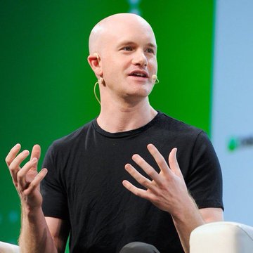 A bald man in a black t-shirt gestures with his hands while speaking, appearing to give a presentation on a stage with a green backdrop.
