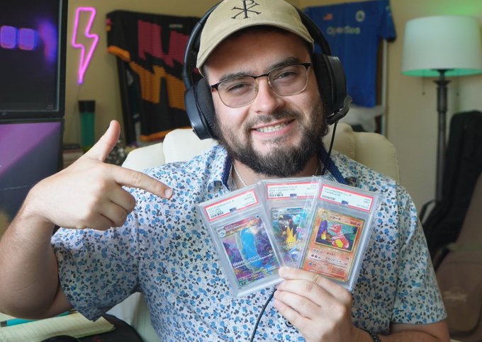 A bearded man wearing glasses, headphones, and a New York Yankees cap holds up three PSA-graded Pokémon cards including what appears to be a Charizard.