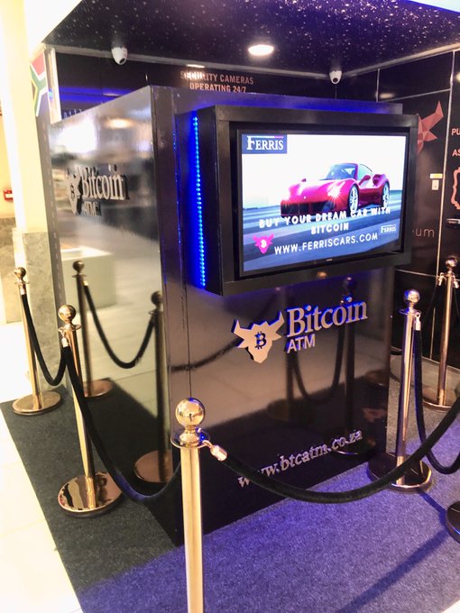 A black Bitcoin ATM branded with 'FERRIS' displayed in a mall or public area, featuring an advertisement on its screen showing a red convertible sports car and the website 'www.ferriscars.com'.