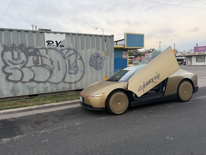 A tan Tesla Cybercab is parked next to a graffiti-covered shipping container, with its signature scissor doors raised and tagged with graffiti that matches the surrounding urban aesthetic. The futuristic vehicle's angular, stealth-bomber-inspired design and solid wheel covers create an aura-farming visual that aligns perfectly with the post's commentary about Tesla's robotaxi intentionally seeking out the most stylish backdrops.