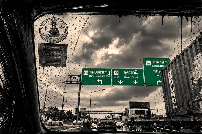 A photograph taken through a rain-streaked window showing green highway signs in Thailand directing toward Nong Bua Lam Phu, Udon Thani, and Sakon Nakhon.