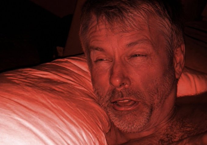 A close-up, red-tinted photo of a shirtless older man with gray hair and a stubble beard lying on a satin pillow.