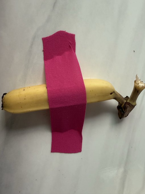 A yellow banana taped to a white wall with pink tape, evoking the infamous $6.2 million banana artwork that crypto entrepreneur Justin Sun famously purchased and ate. The image humorously recreates the conceptual art piece as a visual reference to the viral story.
