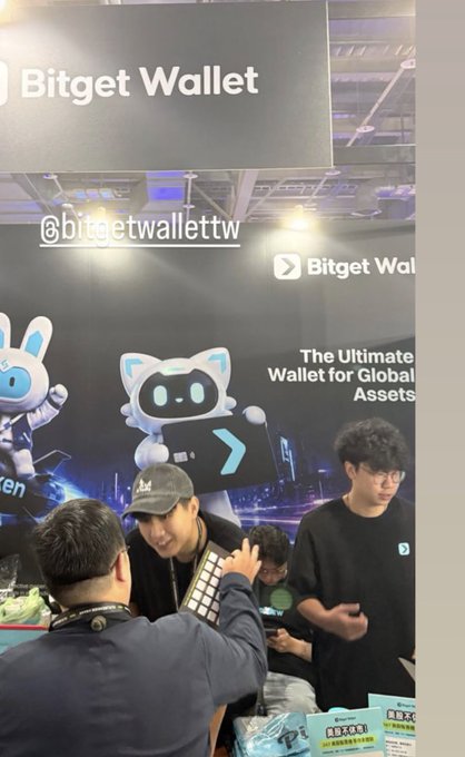 The image shows a busy Bitget Wallet booth at the Taiwan Future Festival, featuring staff members interacting with visitors beneath a banner reading "The Ultimate Wallet for Global Assets," with the @bitgetwallettw handle displayed—capturing the multitasking chaos the poster described as they run their booth, work on hackathon development, and network with friends simultaneously.