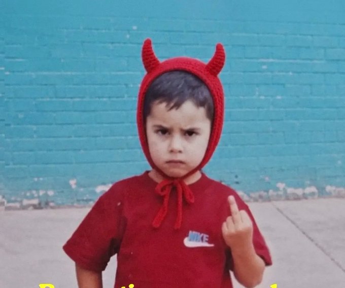 A young boy wearing a red hooded shirt with devil horns and a Nike logo, making an angry face and middle finger gesture against a blue brick wall.
