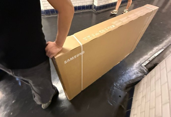 A person in a black shirt and gray pants carries a long, flat Samsung box down a wet subway platform, with another passenger's feet visible in the background. The scene reflects the post's commentary on paying inflated prices ("shelling out obediently") for electronics, likely a Samsung TV, amid burdensome customs fees.