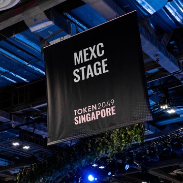 A black hanging banner reading "MEXC STAGE - TOKEN2049 SINGAPORE" is suspended from an industrial ceiling in a convention venue, tying into the #MEXCEventRocks event as part of the TOKEN2049 Singapore crypto conference.