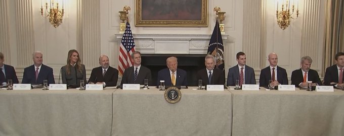 President Donald Trump sits at the center of a long table in the White House, flanked by officials and attendees at the Crypto Summit, with the presidential seal displayed prominently on the tablecloth and the American flag positioned behind him. The setup aligns with the post's announcement of the White House Crypto Summit, showing the gathered leaders seated together for the high-profile discussion on cryptocurrency policy.