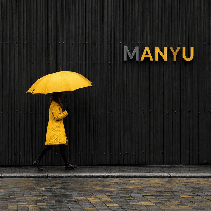 A person wearing a bright yellow raincoat and holding a matching yellow umbrella walks along a sidewalk in front of a dark wooden wall displaying the text 'MANYU'.