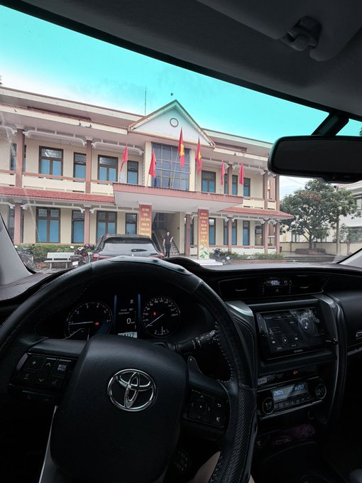 The view from inside a Toyota vehicle shows the steering wheel, dashboard, and center console, looking out through the windshield at a building with Vietnamese flags and a person standing near the entrance.