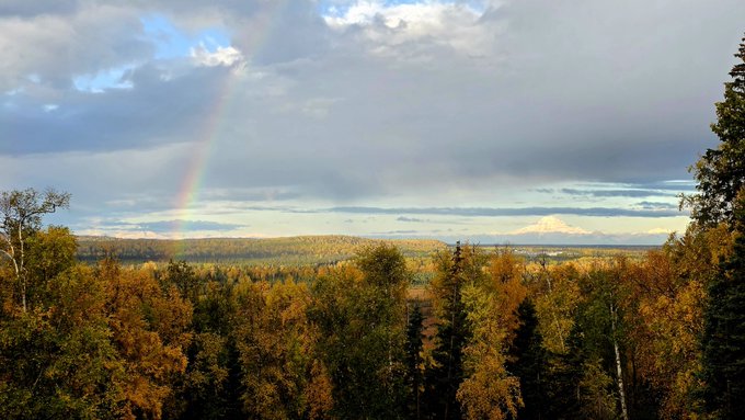Good morning! A vibrant rainbow arcs over Alaska's stunning fall foliage, with golden birches and evergreens stretching toward distant mountain peaks under a partly cloudy sky.