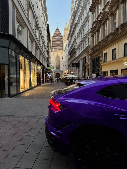 A purple sports car parked on a narrow European street, with a prominent cathedral spire visible in the background.