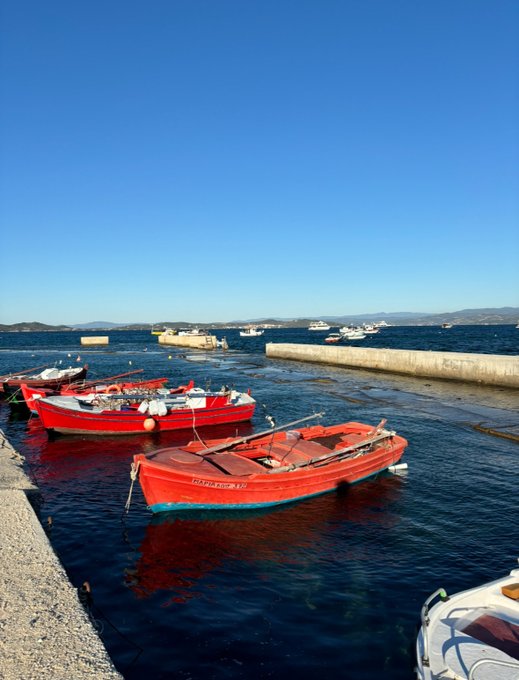 Red fishing boats are docked in a harbor under a clear blue sky, with one boat's name visible as "MARIA ROZ 272". This scene likely relates to the post's mention of a "green port" and "eurosummer" by depicting a peaceful, idyllic setting associated with good fortune and enjoyable times.