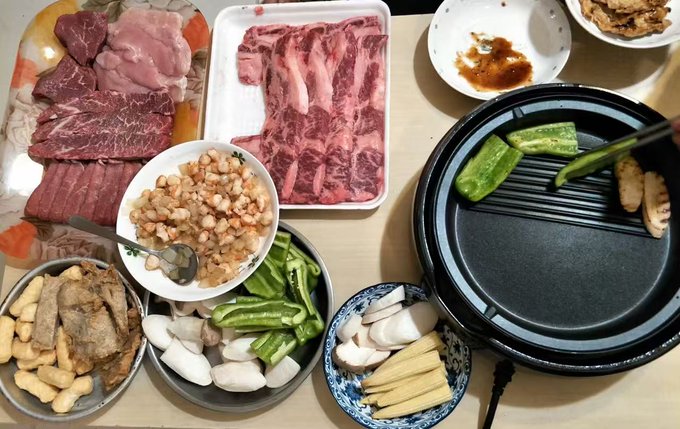 A top-down view of a Korean or Japanese barbecue meal featuring plates of raw beef, pork belly, and assorted vegetables prepared for grilling on a tabletop electric grill.